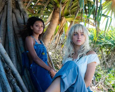 Two women sitting by a large tree in a tropical setting, one in denim overalls and the other in a white blouse.