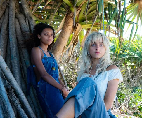 Two women sitting by a large tree in a tropical setting, one in denim overalls and the other in a white blouse.