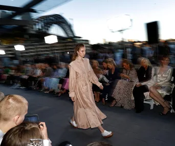 Model walking runway in beige dress at Afterpay Australian Fashion Week 2021 with seated audience and Sydney Harbour Bridge backdrop.