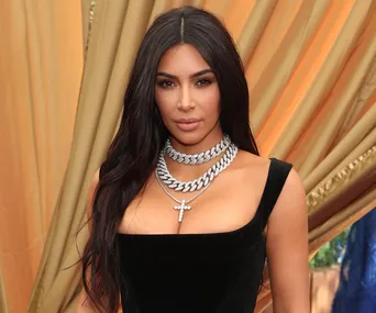 Woman in a black dress with layered silver necklaces, posing against a draped, golden fabric background.