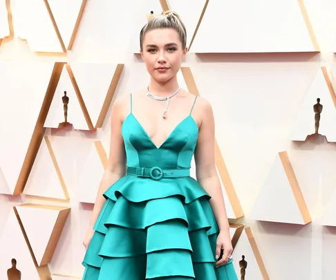 A woman in a teal gown stands on the Oscar red carpet with geometric backdrop patterns.