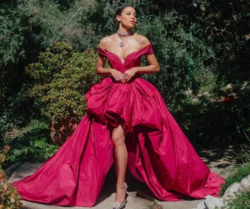 Woman in a vibrant red gown with an elegant necklace, standing outdoors with greenery in the background.