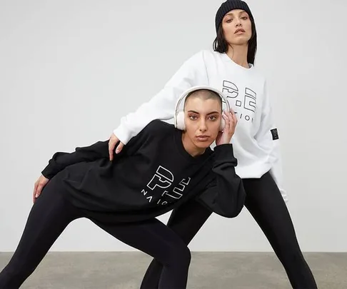 Two models in P.E Nation apparel pose dynamically, one in white, one in black, wearing leggings and beanies.