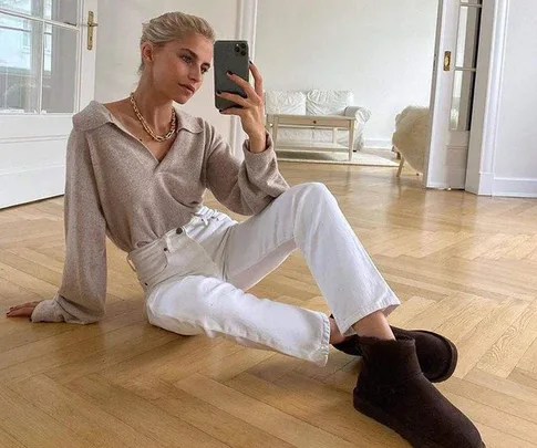 Person taking a mirror selfie wearing a beige sweater, white jeans, and black mini Ugg boots, seated on wooden floor.