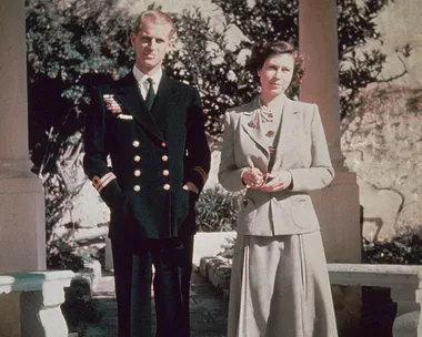 A young man in a navy uniform stands beside a woman in a beige suit, both outdoors in a garden setting.