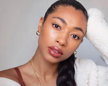 Woman with sleek hairstyle and glowing makeup, wearing hoop earrings and a cozy white sweater, looking at the camera.