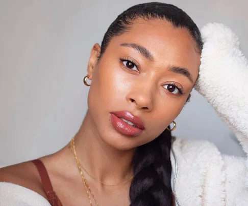 Woman with sleek hairstyle and glowing makeup, wearing hoop earrings and a cozy white sweater, looking at the camera.