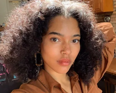 Woman with natural curly hair and nose ring looks directly at camera in a warmly lit room.