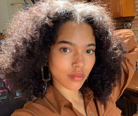 Woman with natural curly hair and nose ring looks directly at camera in a warmly lit room.