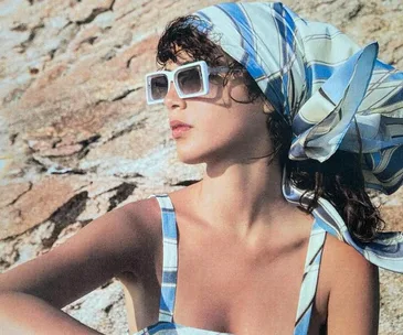 Woman in white square sunglasses and a blue striped headscarf, sitting against a rocky backdrop.