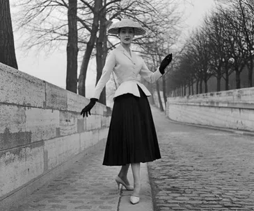 A woman models a Dior Bar Jacket with a full skirt and hat, standing on a tree-lined cobblestone path.