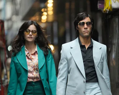 Two characters from "The Serpent" walk confidently in stylish 1970s attire and sunglasses on a city street.