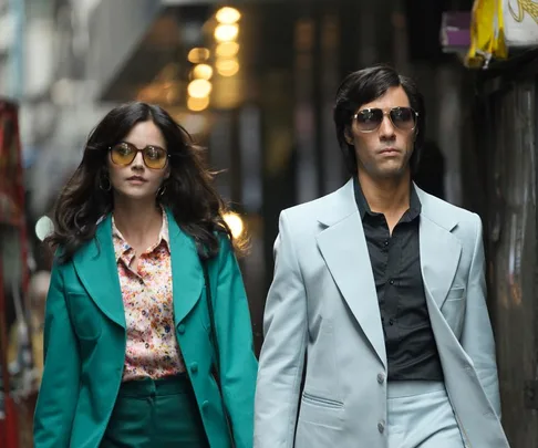 Two characters from "The Serpent" walk confidently in stylish 1970s attire and sunglasses on a city street.