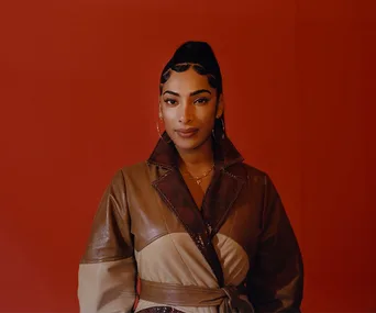 A person in a stylish brown leather jacket stands against a red background, gazing confidently at the camera.