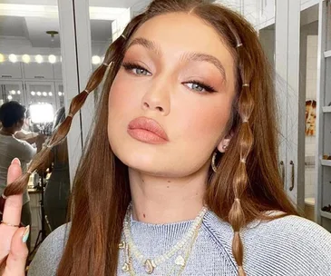 Gigi Hadid with braided hair, wearing a blue top and layered necklaces, posing for a selfie.