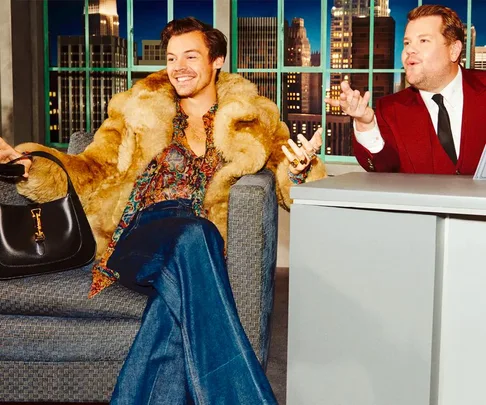 A person wearing a fur coat and flared jeans sits on a couch, holding a handbag, next to another in a suit at a desk.