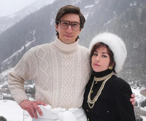 Two people dressed in winter clothing stand outdoors against a snowy mountain backdrop.