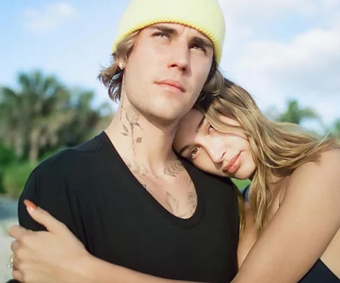 A couple embraces outdoors, with the man wearing a yellow beanie and a black shirt, and the woman resting her head on his shoulder.