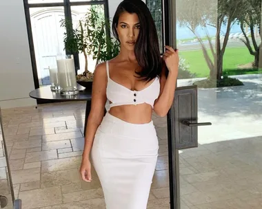 Woman in a white cutout dress standing indoors near a door, with greenery visible outside.