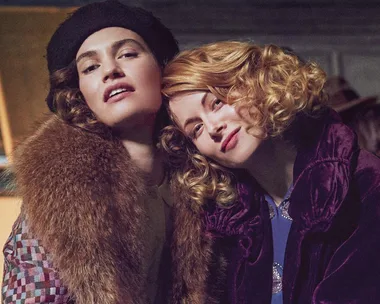 Two women in vintage attire pose closely, one wearing a fur coat and beret, the other in a purple velvet coat with curls.