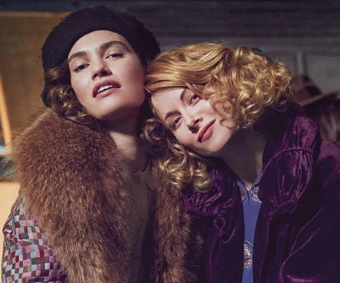 Two women in vintage attire pose closely, one wearing a fur coat and beret, the other in a purple velvet coat with curls.