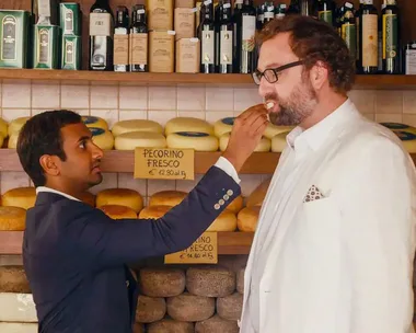 Two men taste cheese in a shop with shelves of cheese wheels in the background.