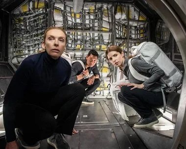 Three astronauts crouch in a spacecraft corridor, looking alert and tense, from the movie "Stowaway."