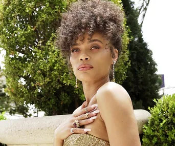 A woman outdoors with curly hair, wearing a gold outfit, jewelry, and looking thoughtfully to the side.