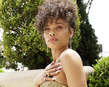 A woman outdoors with curly hair, wearing a gold outfit, jewelry, and looking thoughtfully to the side.