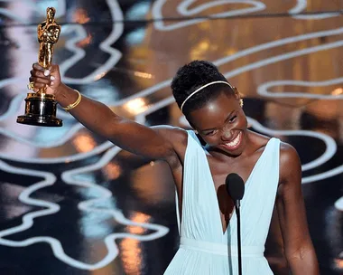 A person in a blue dress holding an Oscar trophy on stage, smiling joyfully.