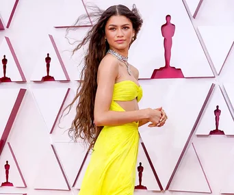 A person in a vibrant yellow dress poses stylishly on a red carpet with geometric patterns in the background.