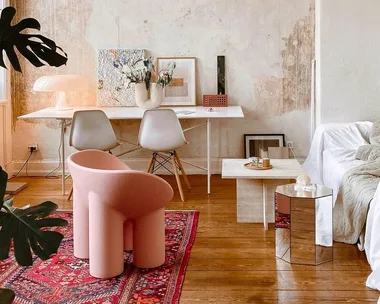 Cozy living room with a pink roly-poly chair, wooden floor, red rug, modern decor, and white desk with art.