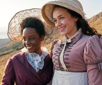 Two women in period costumes smile on a scenic hillside, wearing vintage hats.