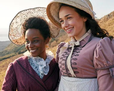 Two women in period costumes smile on a scenic hillside, wearing vintage hats.