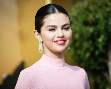 Person in a pink top with diamond earrings, smiling outdoors with greenery in the background.