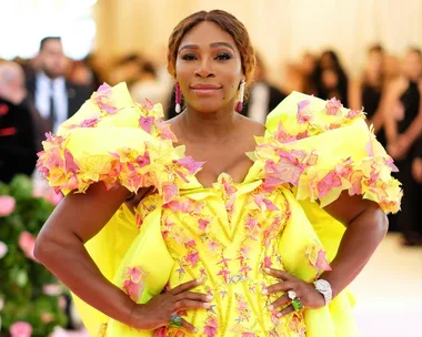 A person wearing a bright yellow floral gown stands confidently with hands on hips at a formal event.