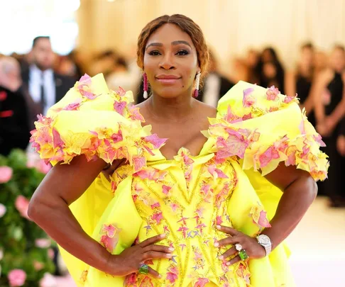 A person wearing a bright yellow floral gown stands confidently with hands on hips at a formal event.