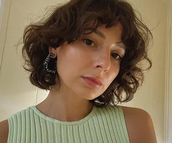 Woman with curly shag haircut and large hoop earrings, wearing a sleeveless mint top, in soft natural lighting.