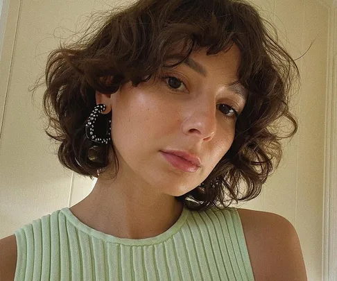 Woman with curly shag haircut and large hoop earrings, wearing a sleeveless mint top, in soft natural lighting.