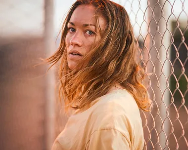 A woman with long hair stands by a chain-link fence, looking back over her shoulder, in dramatic lighting.