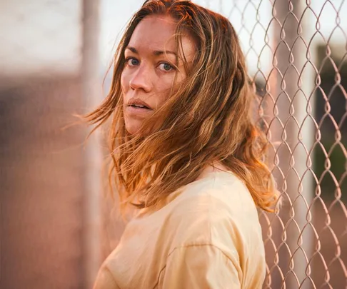 A woman with long hair stands by a chain-link fence, looking back over her shoulder, in dramatic lighting.