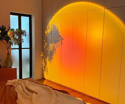 Sunset lamp casts warm, colorful glow with tree shadow on bedroom wall, next to a window and potted plant.