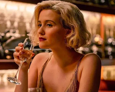 Blonde woman in elegant attire sips wine in a dimly lit bar, scene from "The Girlfriend Experience" season 3.