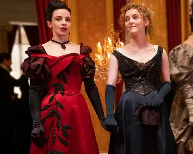 Two women in elegant 19th-century gowns stand together, one in a red dress with black accents, the other in a dark blue dress.