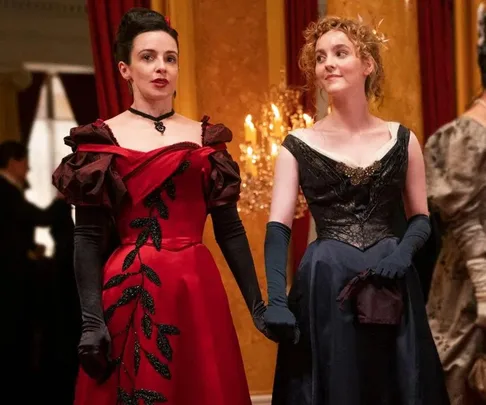 Two women in elegant 19th-century gowns stand together, one in a red dress with black accents, the other in a dark blue dress.