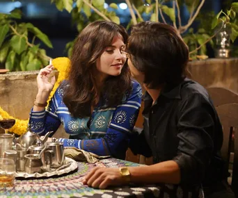 A couple sits close at an outdoor table, with drinks and a teapot, surrounded by plants in a casual setting.