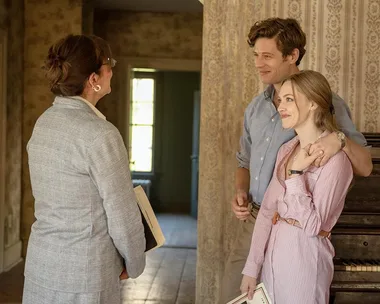 A couple stands smiling in an old, wallpapered room, talking to a woman holding documents.