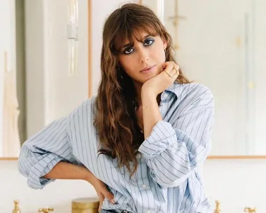 A woman with long hair and bold eye makeup, wearing a striped shirt, poses thoughtfully indoors.