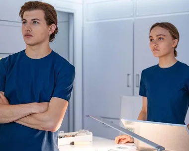 Two individuals in blue uniforms stand in a brightly lit, futuristic room with lab equipment.