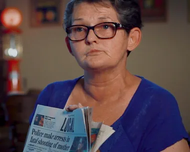 Woman holding a newspaper with headline about arrests in fatal shooting, wearing glasses and a blue shirt.